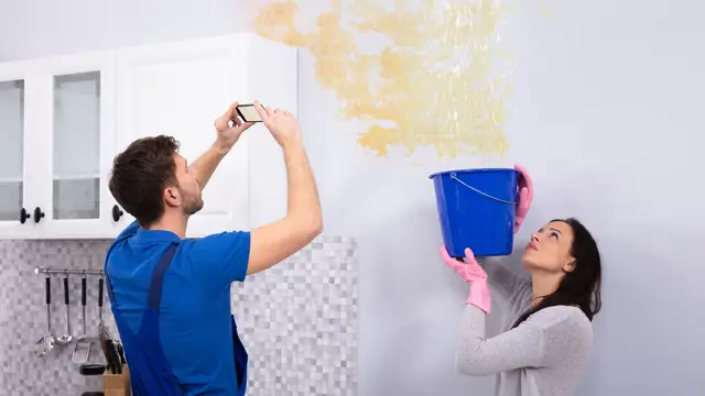Person photographing a water-stained wall while another person holds a bucket